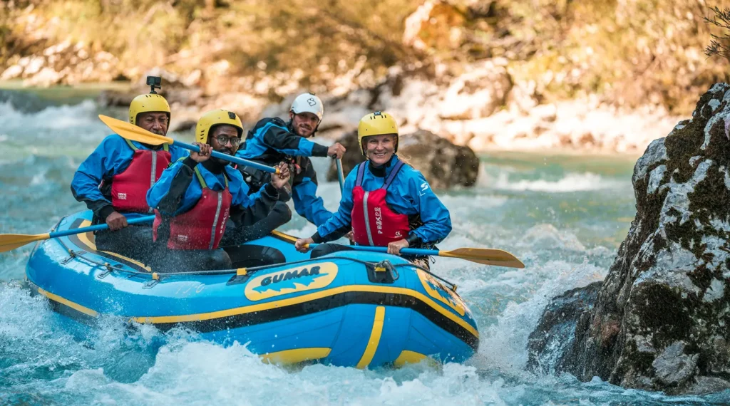 Skupina veslačev v modrem raftu med brzicami smaragdne Soče v Bovcu.