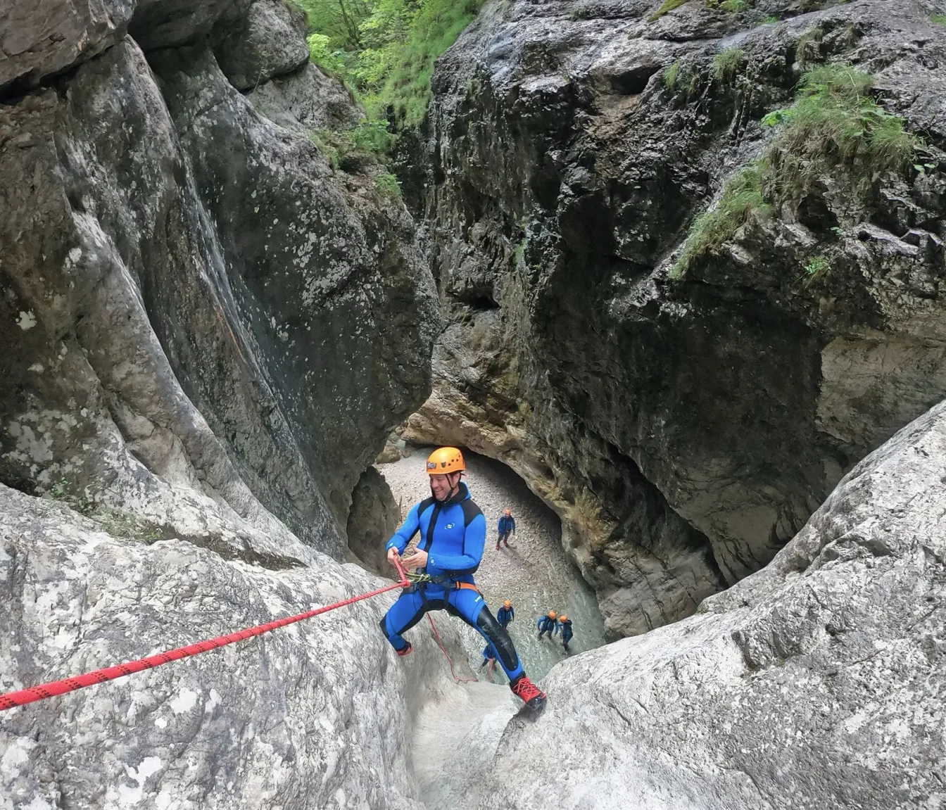 Nasmejan udeleženec se spušča po vrvi po visoki skalni steni, skupina čaka spodaj v kanjonu Rio Ronc.