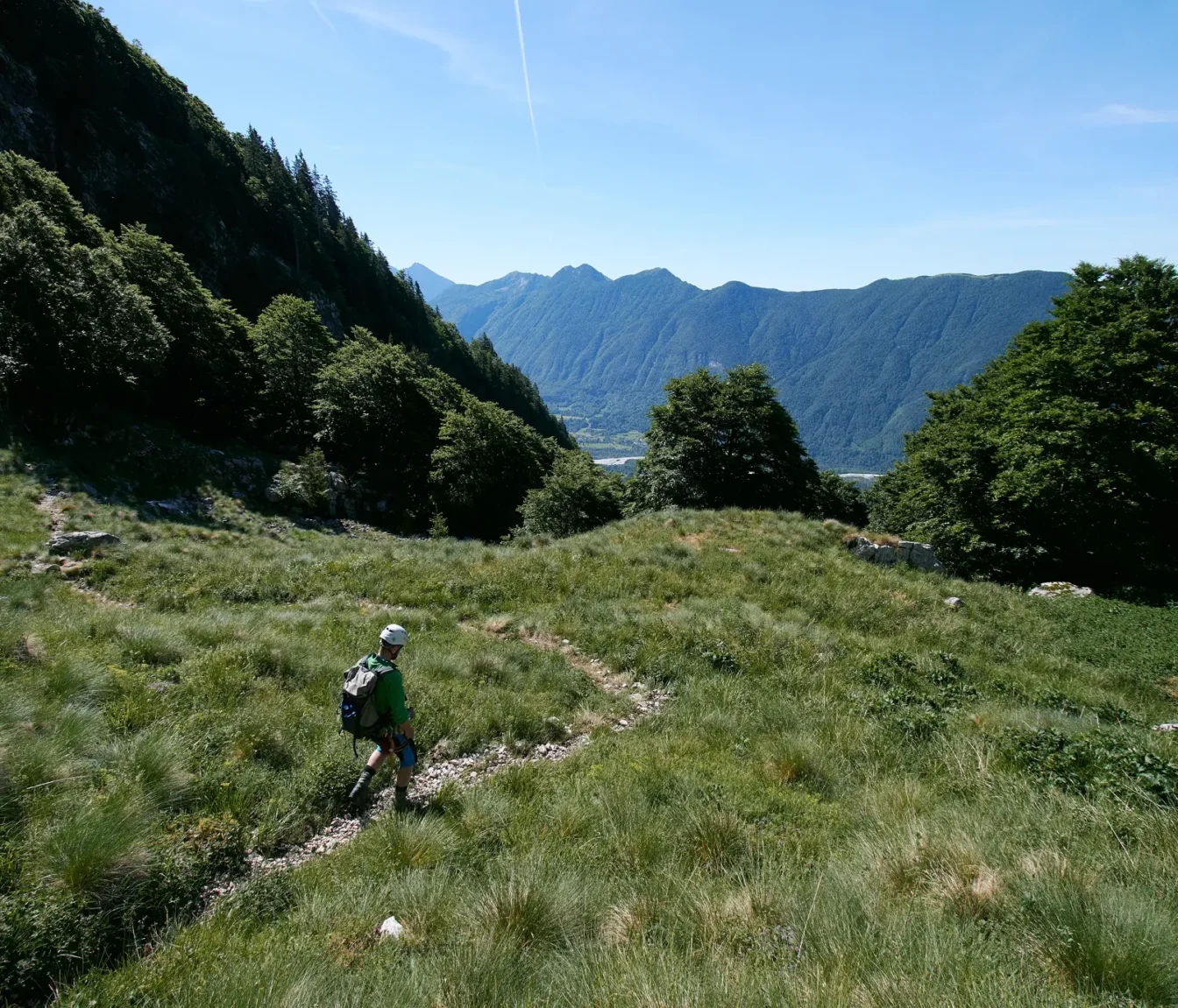 Pohodnik s čelado se spušča po travnati gredi z razgledom na dolino Soče.