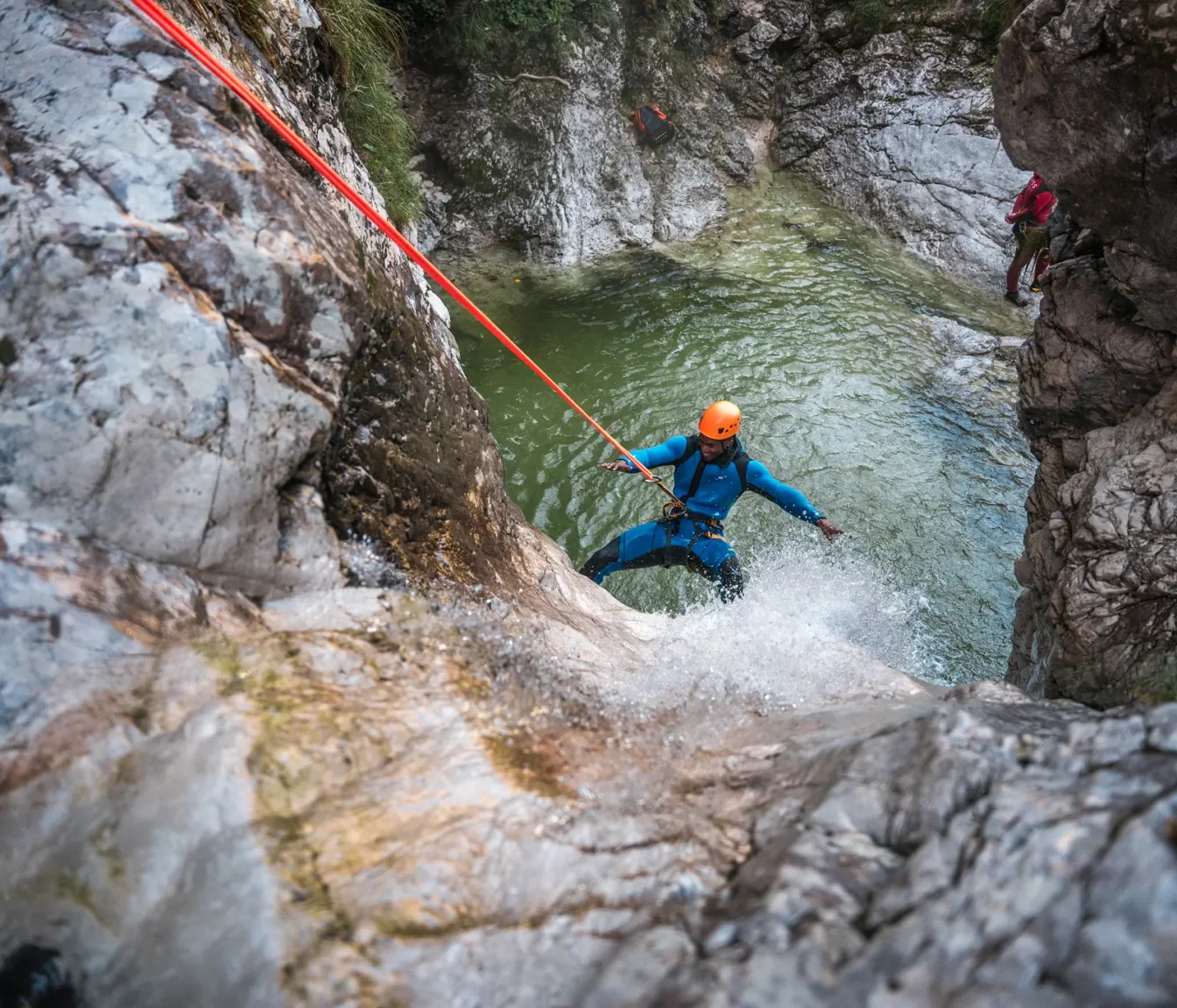 Udeleženec se z oranžno čelado spušča po vrvi nad zelenim tolmunom.